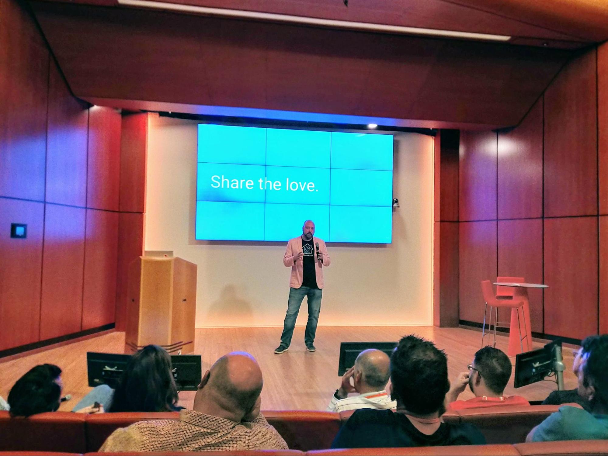 Photo of Paulus Schoutsen speaking to a crowd. The slide behind him shows the text "Share the love".